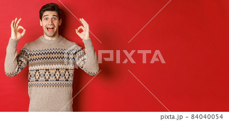 Concept of christmas celebration, winter holidays and lifestyle. Portrait of handsome man in sweater, looking amazed and showing okay signs, guarantee or recommend something good, red background Concept of christmas celebration, winter holidays and lifestyle. Portrait of handsome man in sweater, looking amazed and showing okay signs, guarantee or recommend something good, red background 84040054