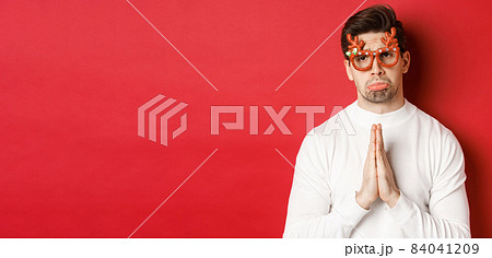 Image of handsome man in christmas party glasses, begging for help or apologizing, need a favour, standing over red background Image of handsome man in christmas party glasses, begging for help or apologizing, need a favour, standing over red background 84041209