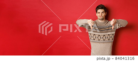 Portrait of skeptical and disappointed handsome man in christmas sweater, dislike new year party, showing thumbs-down, express disapproval, standing over red background 84041316