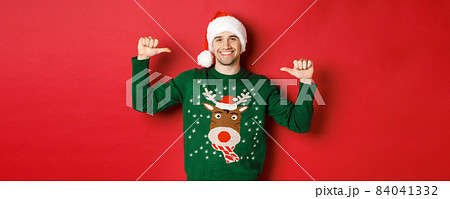 Concept of winter holidays, christmas and lifestyle. Happy attractive guy in santa hat and sweater, pointing at himself with pleased face, standing over red background Concept of winter holidays, christmas and lifestyle. Happy attractive guy in santa hat and sweater, pointing at himself with pleased face, standing over red background 84041332