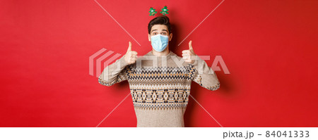 Concept of new year, covid-19 and social distancing. Cheerful caucasian man in sweater and medical mask, celebrating christmas during pandemic, showing thumbs-up, red background Concept of new year, covid-19 and social distancing. Cheerful caucasian man in sweater and medical mask, celebrating christmas during pandemic, showing thumbs-up, red background 84041333