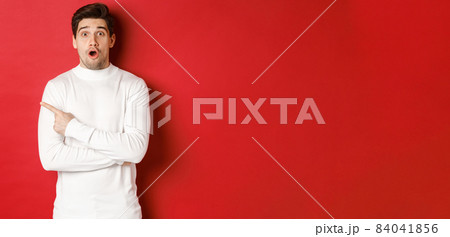 Concept of winter holidays, christmas and lifestyle. Surprised handsome guy pointing finger at new year advertisement, pointing finger left and drop jaw amazed, standing over red background 84041856
