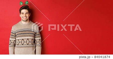 Concept of winter holidays, christmas and celebration. Handsome bearded guy in sweater wishing happy new year, smiling at camera, standing over red background 84041874