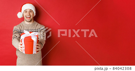 Image of happy man saying merry christmas and giving you a gift in box, smiling cheerful, celebrating winter holidays in santa hat and xmas sweater, standing over red background 84042308