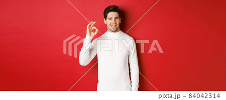 Portrait of charismatic beaded guy in white sweater, assure you, winking and showing okay sign to guarantee good quality, praise excellent choice, standing over red background 84042314