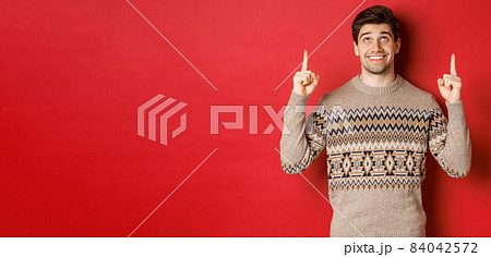 Portrait of dreamy handsome guy thinking about christmas holidays, wearing winter sweater, pointing and looking up at new year promo offer, standing over red background 84042572