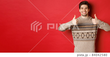 Portrait of happy and pleased handsome guy in christmas sweater, showing thumbs-up and nod in approval, smiling satisfied, standing over red background Portrait of happy and pleased handsome guy in christmas sweater, showing thumbs-up and nod in approval, smiling satisfied, standing over red background 84042580