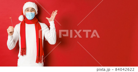 Concept of covid-19, christmas and holidays during pandemic. Cheerful handsome man in medical mask and santa hat, celebrating new year with sparkler, looking excited, red background Concept of covid-19, christmas and holidays during pandemic. Cheerful handsome man in medical mask and santa hat, celebrating new year with sparkler, looking excited, red background 84042622