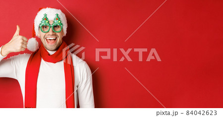 Concept of christmas, winter holidays and celebration. Cheerful guy in santa hat and party glasses, showing thumb-up and smiling pleased, standing over red background 84042623
