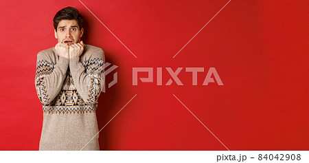 Portrait of scared and timid man in christmas sweater, looking frightened, watching something scary on new year, standing over red background Portrait of scared and timid man in christmas sweater, looking frightened, watching something scary on new year, standing over red background 84042908
