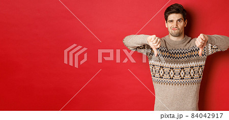 Portrait of skeptical and disappointed handsome man in christmas sweater, dislike new year party, showing thumbs-down, express disapproval, standing over red background Portrait of skeptical and disappointed handsome man in christmas sweater, dislike new year party, showing thumbs-down, express disapproval, standing over red background 84042917