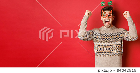 Portrait of thrilled and happy man in party glasses, winter sweater, shouting for joy and raising hands up, winning prize on christmas, achieve new year goal, standing red background Portrait of thrilled and happy man in party glasses, winter sweater, shouting for joy and raising hands up, winning prize on christmas, achieve new year goal, standing red background 84042919