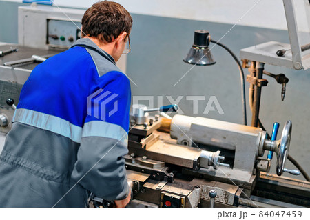 Adult turner of 50-55 years old works in workshop behind lathe. Industrial production of metal parts 84047459