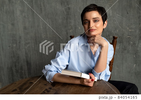 Young brunette woman with short haircut sits at the table in thoughtful pose, holding book in her hand Young brunette woman with short haircut sits at the table in thoughtful pose, holding book in her hand 84054165
