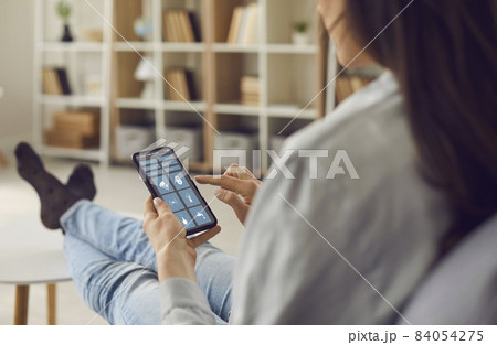 People and technology: Woman sitting in chair, holding mobile phone and using app interface with various icons to control Smart Home system and monitor all rooms in the house, over shoulder close up People and technology: Woman sitting in chair, holding mobile phone and using app interface with various icons to control Smart Home system and monitor all rooms in the house, over shoulder close up 84054275
