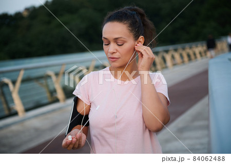 Determined beautiful young athlete sportswoman puts on earphones while standing on the bridge before jogging across the city bridge in the morning on a warm sunny summer day. Sport, fitness concept 84062488