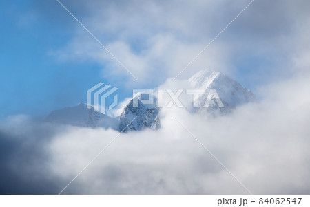 Panoramic alpine sunny landscape with high snowy mountain with peaked top and glacier above low clouds. Big snow covered mountains in sunshine. White-snow pointy peak in sunlight. Panoramic alpine sunny landscape with high snowy mountain with peaked top and glacier above low clouds. Big snow covered mountains in sunshine. White-snow pointy peak in sunlight. 84062547