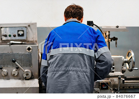 Turner stands behind lathe in production hall and works. View of worker from behind from back in Turner stands behind lathe in production hall and works. View of worker from behind from back in 84076667