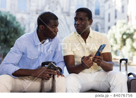 African american travelers sitting on city street, using phones 84076756