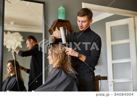 Happy beautiful light hair caucasian woman at beauty salon. Male master is drying her hair 84084004
