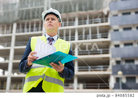 Man architect in helmet with paper documents talking phone at construction site Man architect in helmet with paper documents talking phone at construction site 84085582