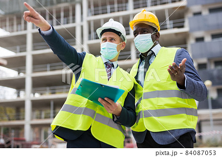 Two engineers in protective masks, discuss a construction plan Two engineers in protective masks, discuss a construction plan 84087053