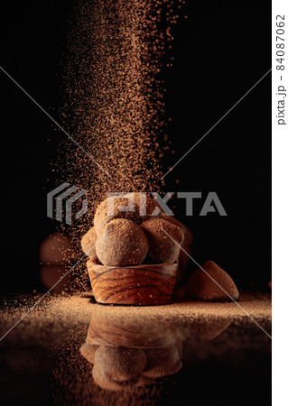 Chocolate truffles in small wooden dish sprinkled with cocoa powder. Black reflective background. 84087062