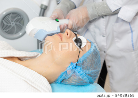 Woman client in a beauty salon is undergoing a photo rejuvenation procedure of alignment skin tone, removing traces of pigmentation. Starting the rejuvenation process by producing own collagen Woman client in a beauty salon is undergoing a photo rejuvenation procedure of alignment skin tone, removing traces of pigmentation. Starting the rejuvenation process by producing own collagen 84089369