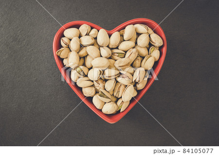 Roasted salted pistachios shelled in heart shaped red bowl on black background, concept of healthy eating vegan food and snack. Close up, selective focus, copy space 84105077