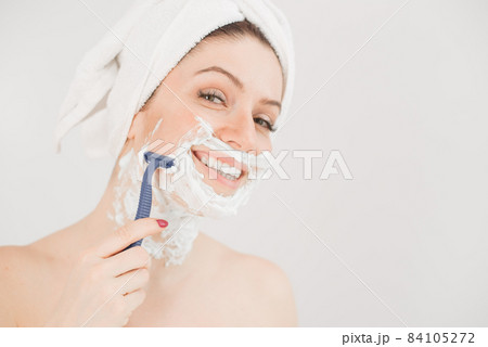 Cheerful caucasian woman with a towel on her head and shaving foam on her face holds a razor on a white background 84105272
