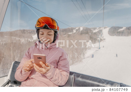 Ski holidays - Woman skier using phone app in gondola ski lift. Girl smiling looking at mobile smartphone wearing ski clothing, helmet and goggles. Ski winter activity vacation concept Ski holidays - Woman skier using phone app in gondola ski lift. Girl smiling looking at mobile smartphone wearing ski clothing, helmet and goggles. Ski winter activity vacation concept 84107749