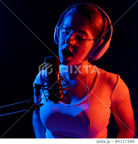 Caucasian woman in glasses and headphones sings into a microphone in neon light on a black background. An emotional girl is recording a song in a recording studio Caucasian woman in glasses and headphones sings into a microphone in neon light on a black background. An emotional girl is recording a song in a recording studio 84117380