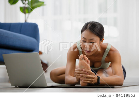 In the morning, an Asian woman is studying online yoga class, standing forward bend exercise, head to knees, uttanasana stance. In the morning, an Asian woman is studying online yoga class, standing forward bend exercise, head to knees, uttanasana stance. 84119952