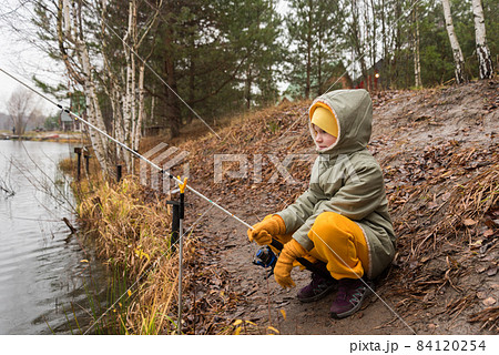 The child is a fisherman by the autumn lake. A child in warm clothes in the rain sits on the shore with a fishing rod The child is a fisherman by the autumn lake. A child in warm clothes in the rain sits on the shore with a fishing rod 84120254