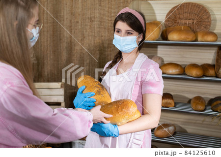 A customer buys more loaves of bread for her family. The saleswoman serves the bread. A saleswoman during the COVID-19 pandemic is wearing gloves and a face mask at a bakery store. A customer buys more loaves of bread for her family. The saleswoman serves the bread. A saleswoman during the COVID-19 pandemic is wearing gloves and a face mask at a bakery store. 84125610