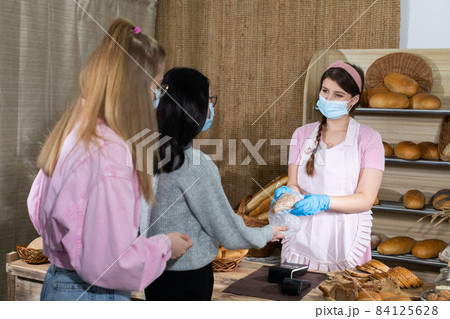 A saleswoman in a protective mask serves bought bread by two teenagers. Sales during the ongoing COVID-19 pandemic. A saleswoman in a protective mask serves bought bread by two teenagers. Sales during the ongoing COVID-19 pandemic. 84125628
