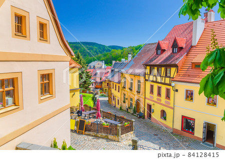 Loket town narrow street, colorful traditional typical buildings with multicolored walls, cobblestone road, wooden fence, green hills background, Karlovy Vary Region, West Bohemia, Czech Republic 84128415