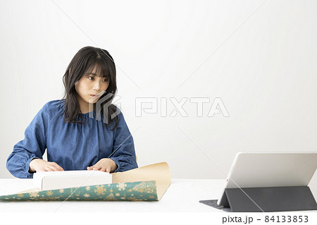 ネットの動画を見ながら机でプレゼントの梱包をする若い女性 84133853