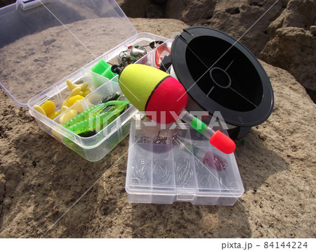 open boxes of translucent plastic with fishing tackle floats and a reel of line lie on a large stone. open boxes of translucent plastic with fishing tackle floats and a reel of line lie on a large stone. 84144224