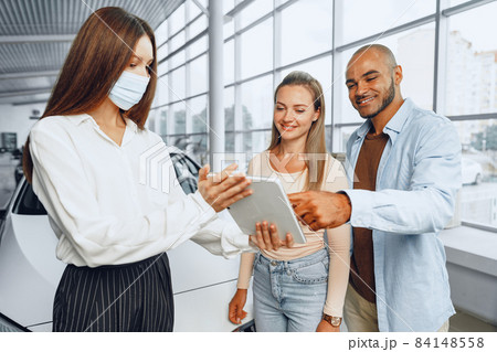 Car saleswoman wearing medical mask shows buyers couple something on digital tablet 84148558