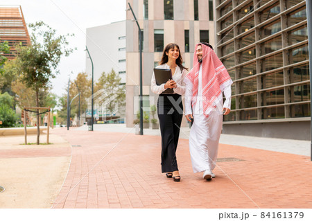 Smiling arab businessman in traditional clothes walking city street with his young attractive european female assistant, saudi man client talking with caucasian businesswoman before signing contract Smiling arab businessman in traditional clothes walking city street with his young attractive european female assistant, saudi man client talking with caucasian businesswoman before signing contract 84161379