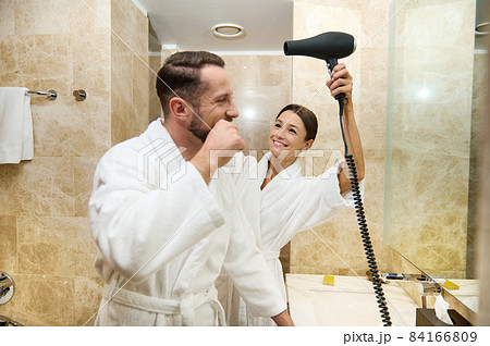 Happy middle aged loving couple having fun together at morning hygiene routine in bathroom. Handsome guy brushing teeth, smiles while his wife or girlfriend drying his hair using hair dryer 84166809