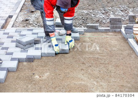 A worker wearing protective gloves carefully lays paving slabs along a stretched thread on a prepared flat sandy area. Copy space. A worker wearing protective gloves carefully lays paving slabs along a stretched thread on a prepared flat sandy area. Copy space. 84170304