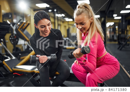 Female Coach shows the client an exercise for biceps with dumbbell while sitting on the bench in gym Female Coach shows the client an exercise for biceps with dumbbell while sitting on the bench in gym 84171380