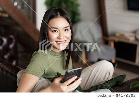 Smiling asian woman sitting on couch at home in cozy clothes, using mobile phone application, looking happy at camera 84173585