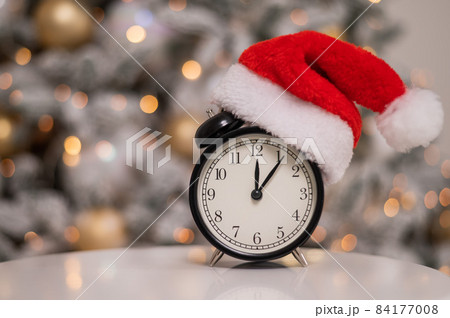 Alarm clock in a santa hat against the background of a christmas tree. Alarm clock in a santa hat against the background of a christmas tree. 84177008
