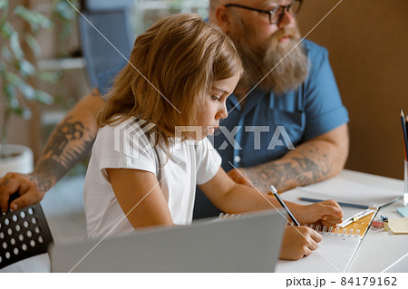Pretty little girl writes in copybook sitting nera laptop doing homework with daddy at home Pretty little girl writes in copybook sitting nera laptop doing homework with daddy at home 84179162