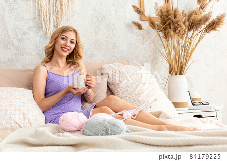 Young woman holding a mug with tea or coffee sitting on the bed in the bedroom. Near lie yarn and knitting needles. 84179225