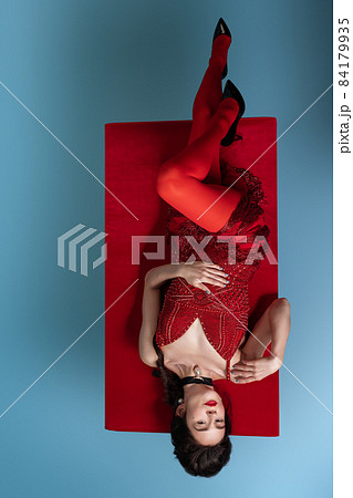 Top view of young adorable woman in dark red, wine color evening dress lying on table with red cloth isolated on blue background. 84179935