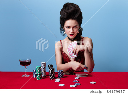 One young beautiful gorgeous woman in dark red, wine color evening dress makes bets, plays poker isolated on blue background. 84179957